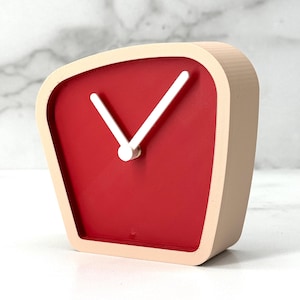 Puede incluir: Un reloj de escritorio moderno, de forma cuadrada, con esfera roja y manecillas blancas. El reloj tiene un marco beige claro y un diseño minimalista. El reloj es un elemento decorativo para el hogar o la oficina.