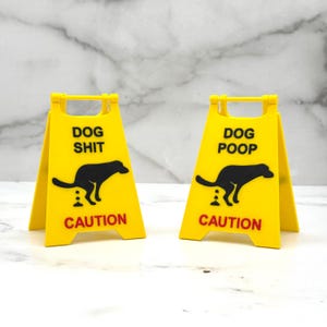 May include: Two yellow caution signs with black dog silhouettes and the words "DOG SHIT" and "DOG POOP" in black text. The word "CAUTION" is in red text. The signs are designed to warn of dog waste.
