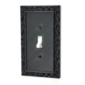 Può includere: Copri interruttore nero con una cornice decorativa e ornata. L'interruttore è bianco e la copertura è montata su una parete bianca. La cornice presenta un design dettagliato a volute.