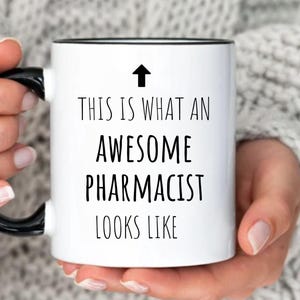 May include: White ceramic coffee mug with a black rim and handle. The mug has black text that reads "This is what an awesome pharmacist looks like" with an arrow pointing upwards.