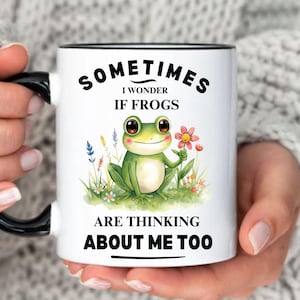 May include: White ceramic mug with a black handle and rim, featuring a green frog illustration holding a flower. Text reads "Sometimes I wonder if frogs are thinking about me too." Additional text: "More Color Options" and "We Accept Custom Requests."