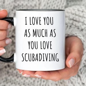 May include: White ceramic mug with a black handle and rim, featuring the text "I LOVE YOU AS MUCH AS YOU LOVE SCUBADIVING" in black. The mug also has the text "MORE COLOR OPTIONS" and "WE ACCEPT CUSTOM REQUESTS".
