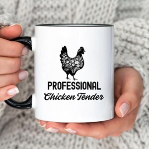 May include: White ceramic mug with a black rim and handle. The mug features a black and white illustration of a chicken with the text "Professional Chicken Tender".