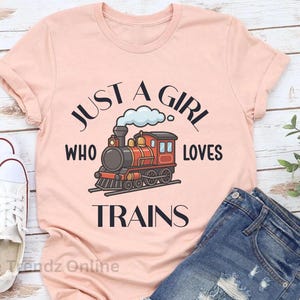 May include: A heather peach short-sleeve t-shirt featuring a graphic of a red and black steam locomotive with a cloud of smoke. The text printed in black reads, "JUST A GIRL WHO LOVES TRAINS," arranged in a circular pattern around the train illustration.