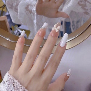 May include: White and nude long false nails with a white tip and a small white bow on the ring finger. The nails are decorated with rhinestones.