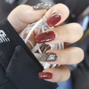 May include: Close-up of a hand with red nail polish and a single nail with a silver rhinestone design.