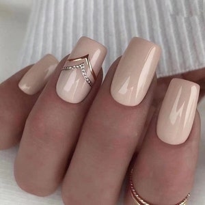 May include: Close-up of a hand with nude-colored square-shaped nails. The middle fingernail has a gold and rhinestone design in the shape of a chevron.