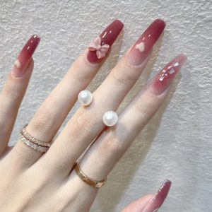 Könnte beinhalten: Ein Handmodell trägt einen Satz langer, ombrefarbener pinkfarbener und weißer Press-on-Nägel mit verschiedenen Verzierungen, darunter eine rosa Schleife, ein weißes Herz und weißer Glitzer. Die Hand trägt auch zwei goldene Ringe mit weißen Perlenakzenten.