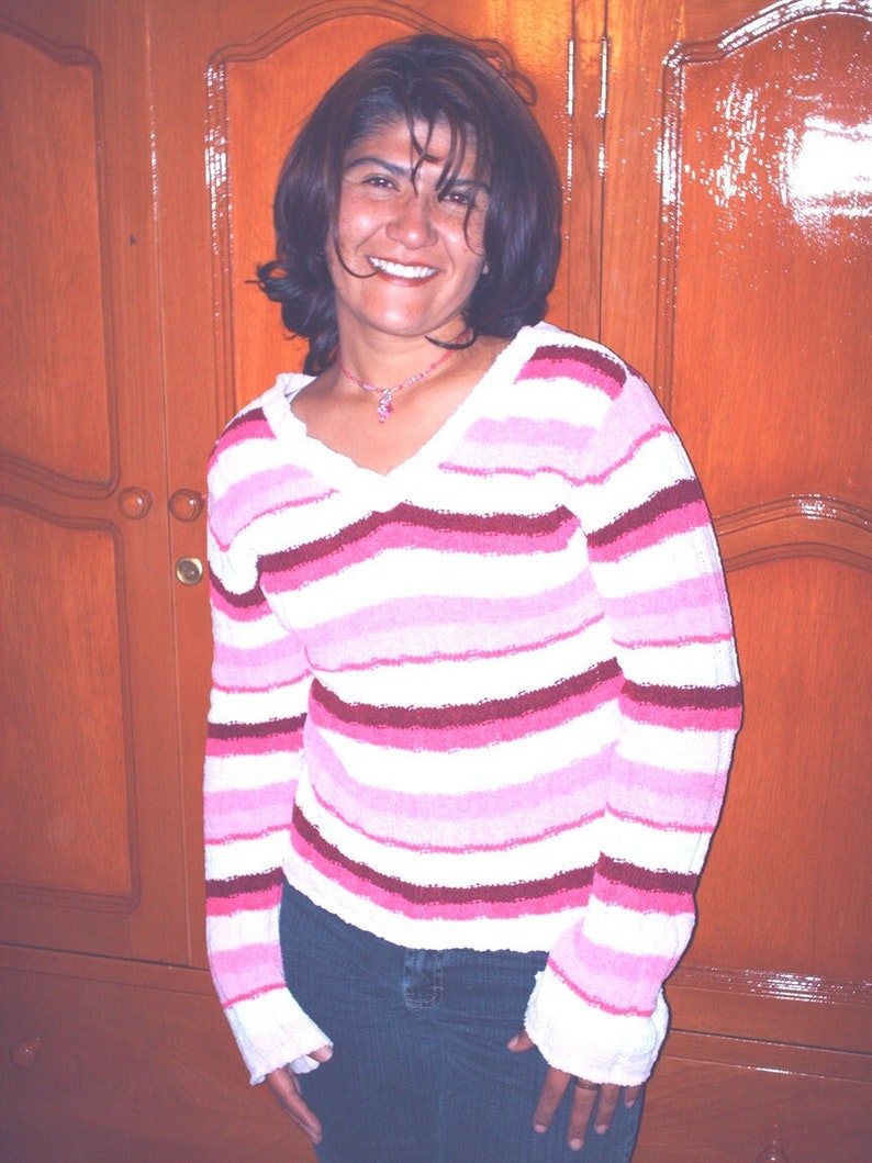Pode incluir: Uma mulher com cabelo escuro est&aacute; vestindo um su&eacute;ter listrado de rosa e branco com mangas compridas. Ela est&aacute; sorrindo e olhando para a c&acirc;mera. Ela est&aacute; vestindo jeans azul e um colar de prata.