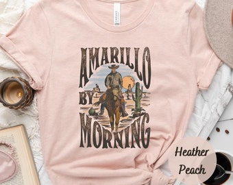 May include: A pink t-shirt with the text "Amarillo by Morning" in a vintage style font. The design features a cowboy riding a horse in a desert landscape. The shirt is made of a soft, comfortable fabric and is perfect for everyday wear.