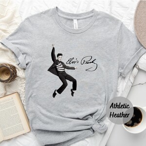 Op de afbeelding: Een grijs T-shirt met een zwart-wit afbeelding van Elvis Presley in een jumpsuit en zijn handtekening. De tekst "Athletic Heather" is gedrukt in de rechterbenedenhoek van het shirt.