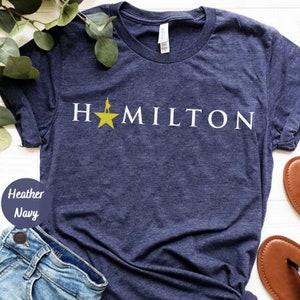 May include: A heather navy blue t-shirt with the word "HAMILTON" printed in white with a yellow star and a silhouette of a person standing on the star.