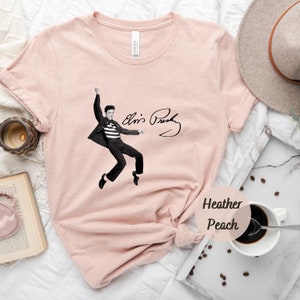 Op de afbeelding: Een roze T-shirt met een zwart-witte afbeelding van Elvis Presley in een sprongpose met zijn handtekening eronder. De tekst "Heather Peach" staat in een cirkel in de rechterbenedenhoek van de afbeelding.