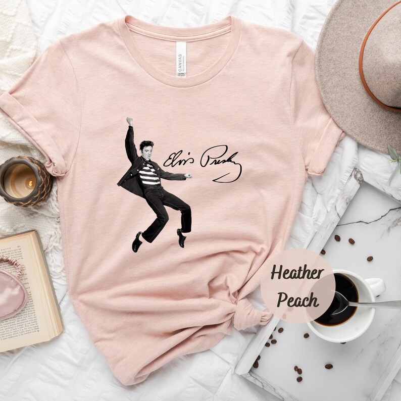 Op de afbeelding: Een roze T-shirt met een zwart-witte afbeelding van Elvis Presley in een sprongpose met zijn handtekening eronder. De tekst "Heather Peach" staat in een cirkel in de rechterbenedenhoek van de afbeelding.