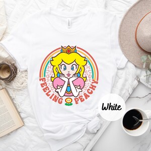 May include: White t-shirt with a retro-style graphic of Princess Peach from the Super Mario Bros. video game series. The graphic features Peach with a crown, a pink dress, and a flower. The text "Feeling Peachy" is written around the graphic.