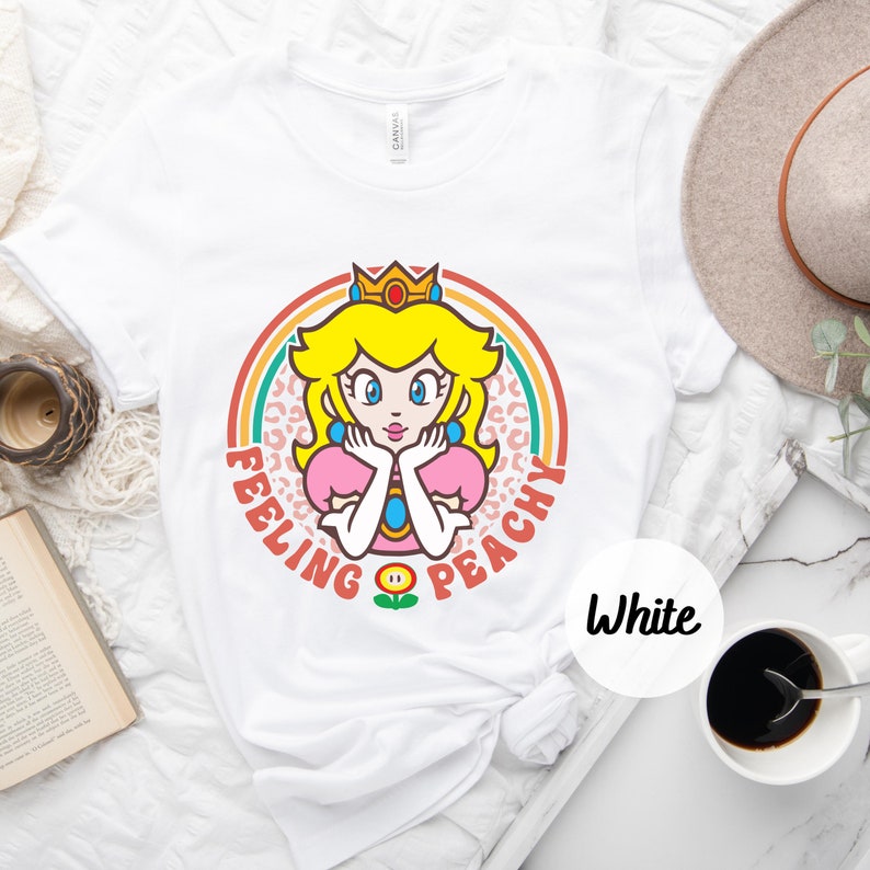 May include: White t-shirt with a retro-style graphic of Princess Peach from the Super Mario Bros. video game series. The graphic features Peach with a crown, a pink dress, and a flower. The text "Feeling Peachy" is written around the graphic.