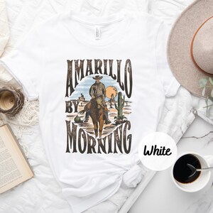 Op de afbeelding: Wit T-shirt met een vintage grafisch ontwerp met een cowboy die op een paard rijdt in een woestijnlandschap. De tekst "Amarillo By Morning" is gedrukt in bruine letters.