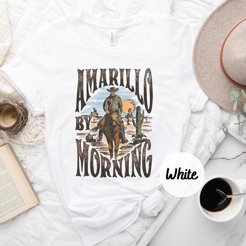 Op de afbeelding: Wit T-shirt met een vintage grafisch ontwerp met een cowboy die op een paard rijdt in een woestijnlandschap. De tekst "Amarillo By Morning" is gedrukt in bruine letters.
