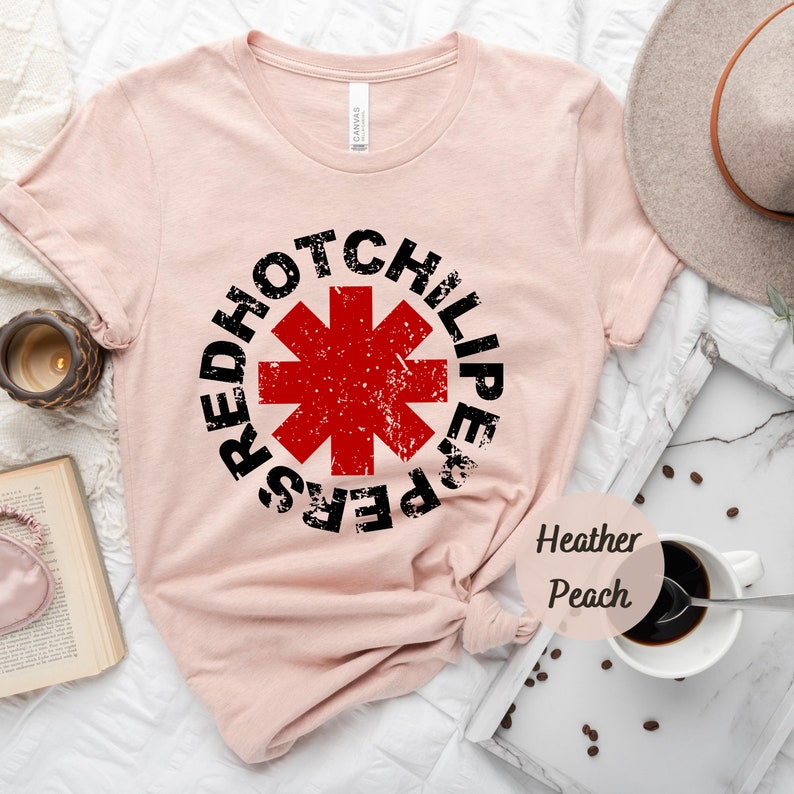 May include: A pink t-shirt with a black and red graphic of the Red Hot Chili Peppers logo. The logo is a red star with a black outline and the words "Red Hot Chili Peppers" surrounding it. The shirt is made of a soft, comfortable material and is perfect for everyday wear.