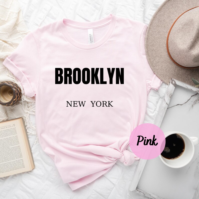 May include: Pink t-shirt with the text "BROOKLYN NEW YORK" in black lettering.