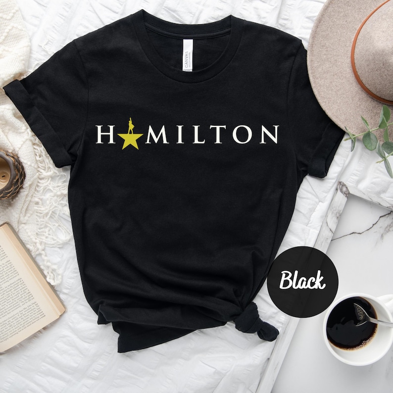 Op de afbeelding: Zwart T-shirt met een witte ster en de tekst "HAMILTON" gedrukt op de voorkant.
