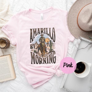 Op de afbeelding: Roze T-shirt met een vintage grafisch ontwerp met een cowboy te paard met de tekst "Amarillo By Morning".