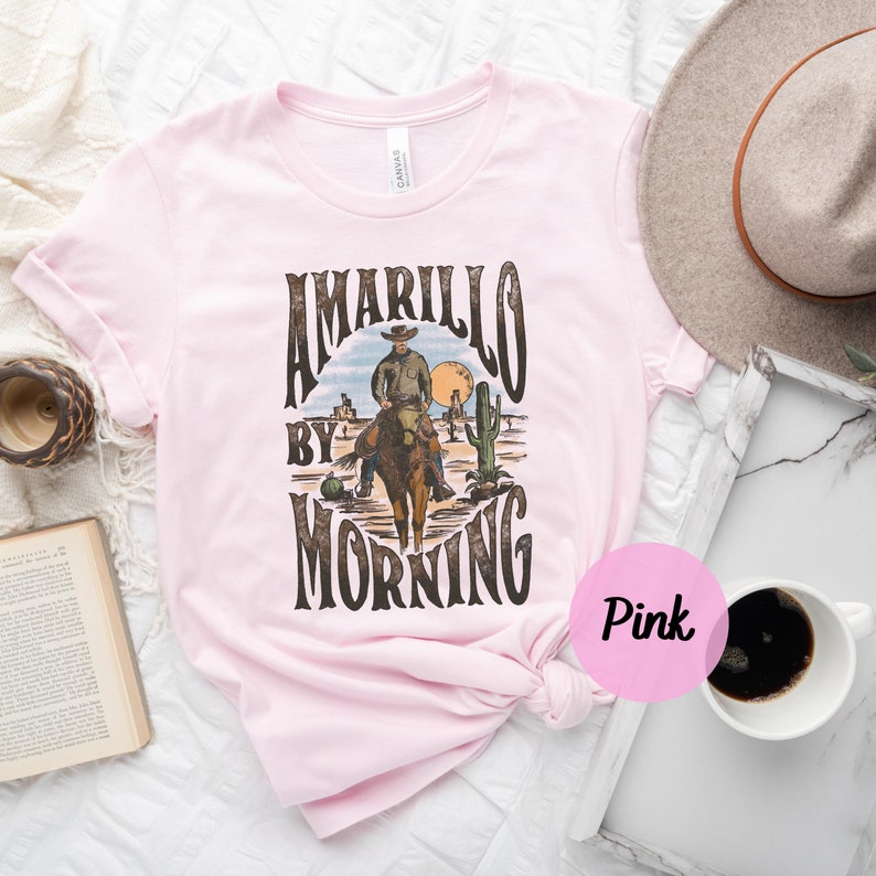 Op de afbeelding: Roze T-shirt met een vintage grafisch ontwerp met een cowboy te paard met de tekst "Amarillo By Morning".