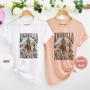 Op de afbeelding: Twee T-shirts met een vintage grafisch ontwerp van een cowboy die op een paard rijdt voor een woestijnlandschap. De tekst op de shirts luidt "Amarillo By Morning". Een shirt is wit en het andere is heidekleurig.