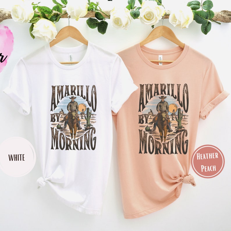 Op de afbeelding: Twee T-shirts met een vintage grafisch ontwerp van een cowboy die op een paard rijdt voor een woestijnlandschap. De tekst op de shirts luidt "Amarillo By Morning". Een shirt is wit en het andere is heidekleurig.