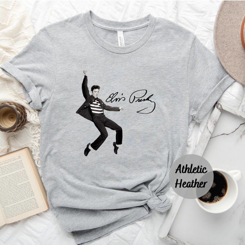 Op de afbeelding: Een grijs T-shirt met een zwart-wit afbeelding van Elvis Presley in een jumpsuit en zijn handtekening. De tekst "Athletic Heather" is gedrukt in de rechterbenedenhoek van het shirt.