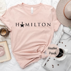 Op de afbeelding: Een lichtroze T-shirt met het woord "HAMILTON" in zwart gedrukt, met een silhouet van een persoon die op een ster staat. Het T-shirt ligt op een wit oppervlak met een kopje koffie en koffiebonen.