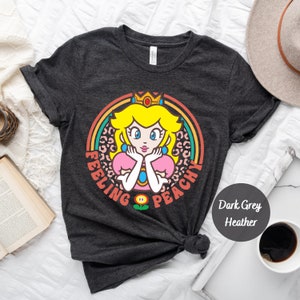 May include: Dark grey heather t-shirt with a retro-style graphic of Princess Peach from the Super Mario Bros. video game series. The graphic features Peach with a crown, a pink dress, and a leopard print background. The text "Feeling Peachy" is written around the graphic.