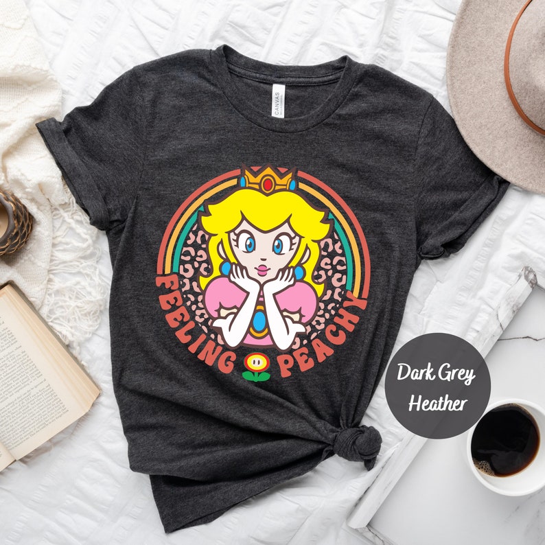 May include: Dark grey heather t-shirt with a retro-style graphic of Princess Peach from the Super Mario Bros. video game series. The graphic features Peach with a crown, a pink dress, and a leopard print background. The text "Feeling Peachy" is written around the graphic.