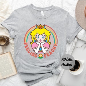 May include: A gray t-shirt with a retro rainbow design featuring Princess Peach from the Super Mario Bros. video game series. The text "Feeling Peachy" is printed on the shirt.