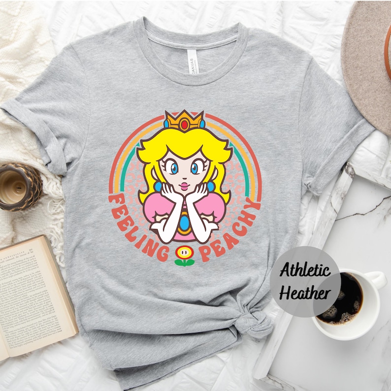 May include: A gray t-shirt with a retro rainbow design featuring Princess Peach from the Super Mario Bros. video game series. The text "Feeling Peachy" is printed on the shirt.