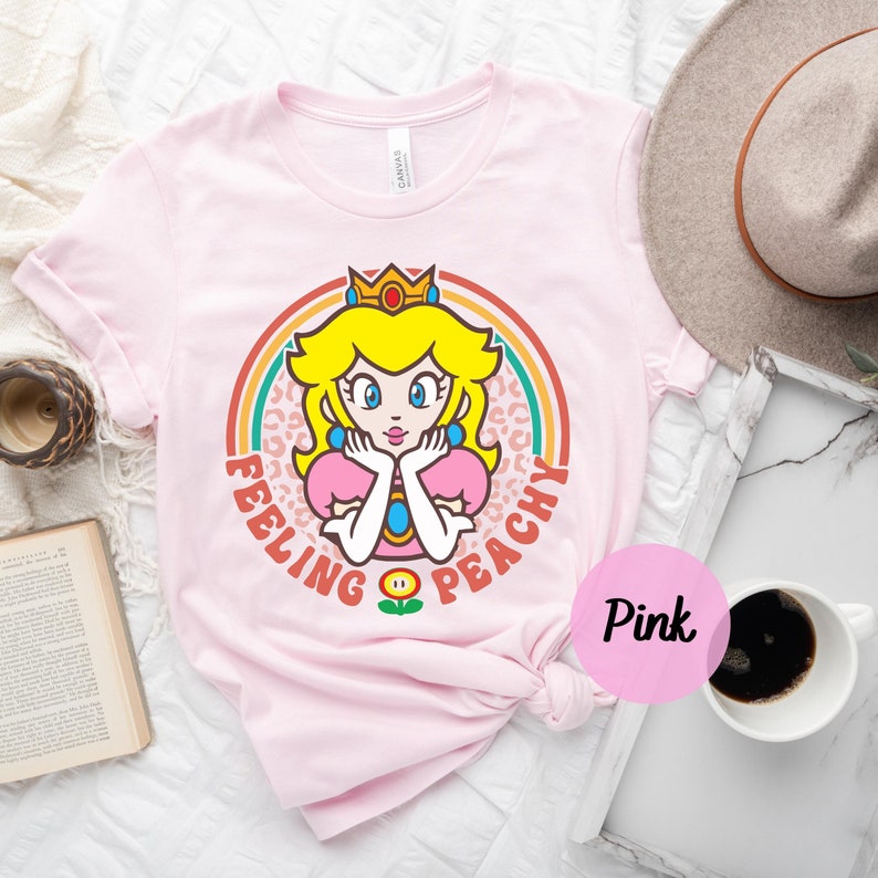 May include: Pink t-shirt with a graphic of Princess Peach from Super Mario Bros. The graphic features Peach with a crown, a pink dress, and a rainbow behind her. The text "Feeling Peachy" is written around the graphic.