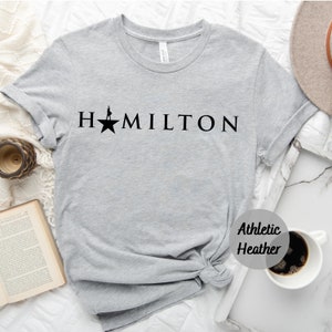 Op de afbeelding: Een grijs T-shirt met het woord "HAMILTON" in zwart gedrukt, met een ster en een silhouet van een persoon die op de ster staat. Het T-shirt is gemarkeerd met "Athletic Heather".