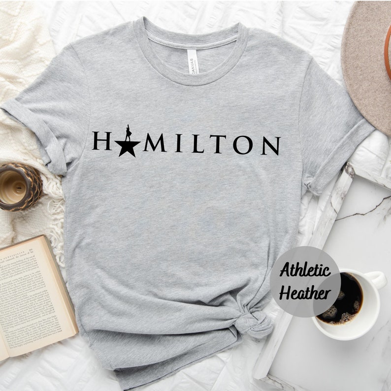 Op de afbeelding: Een grijs T-shirt met het woord "HAMILTON" in zwart gedrukt, met een ster en een silhouet van een persoon die op de ster staat. Het T-shirt is gemarkeerd met "Athletic Heather".
