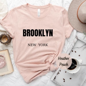 May include: A light pink t-shirt with the text "BROOKLYN NEW YORK" printed in black. The shirt is made of a soft, comfortable material and has a relaxed fit. The shirt is photographed on a white background with a coffee cup, coffee beans, and a book.