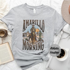 Op de afbeelding: Een grijs T-shirt met een vintage grafisch ontwerp met een cowboy die op een paard rijdt met de tekst "Amarillo by Morning". Het shirt is gemaakt van atletisch heather-stof.