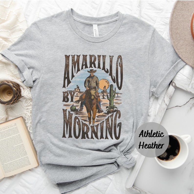 Op de afbeelding: Een grijs T-shirt met een vintage grafisch ontwerp met een cowboy die op een paard rijdt met de tekst "Amarillo by Morning". Het shirt is gemaakt van atletisch heather-stof.