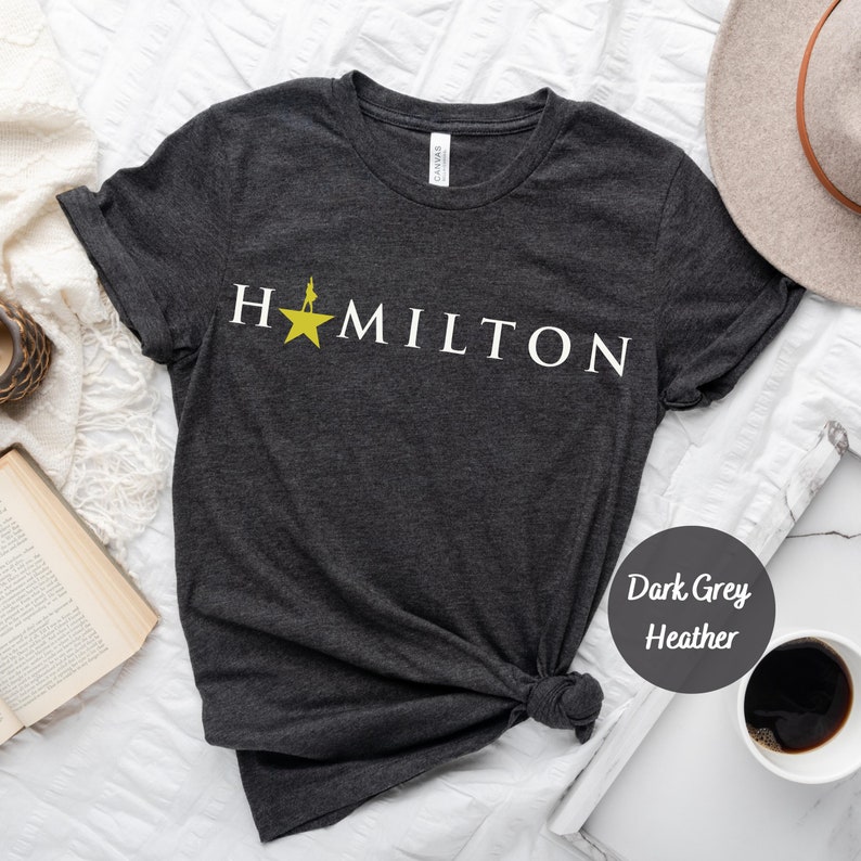 Op de afbeelding: Donkergrijs heather t-shirt met een witte afbeelding van een ster en het woord "HAMILTON".
