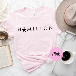 Op de afbeelding: Roze T-shirt met een zwarte ster en het woord "HAMILTON" gedrukt op de voorkant.