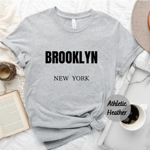 May include: Gray t-shirt with black text that reads "BROOKLYN NEW YORK" in a simple, modern font. The shirt is made of a soft, comfortable material and has a relaxed fit.