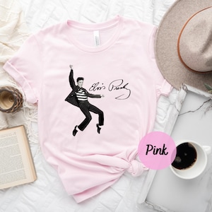 Op de afbeelding: Roze T-shirt met een zwart-witte afbeelding van Elvis Presley die optreedt met zijn handtekening eronder. De tekst luidt "Elvis Presley".