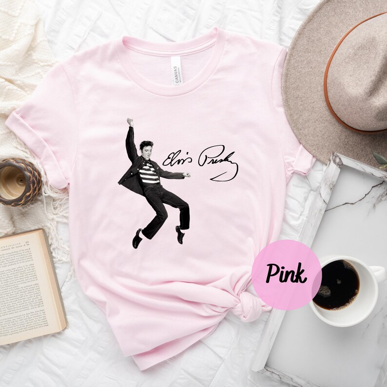 Op de afbeelding: Roze T-shirt met een zwart-witte afbeelding van Elvis Presley die optreedt met zijn handtekening eronder. De tekst luidt "Elvis Presley".