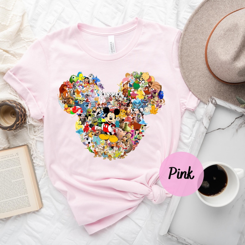May include: Pink t-shirt with a Mickey Mouse head design made up of various Disney characters. The shirt is on a white surface with a brown hat and a cup of coffee.
