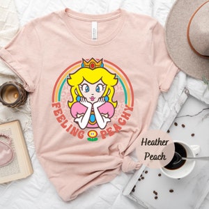 May include: A pink t-shirt with a retro-style graphic of Princess Peach from the Super Mario Bros. video game series. The graphic features Princess Peach with a crown, a rainbow, and the text "Feeling Peachy".