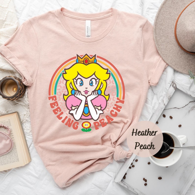 May include: A pink t-shirt with a retro-style graphic of Princess Peach from the Super Mario Bros. video game series. The graphic features Princess Peach with a crown, a rainbow, and the text "Feeling Peachy".