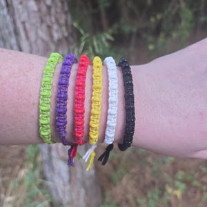 May include: A set of six woven cord bracelets in neon green, purple, red, yellow, white, and black. Each bracelet features a repeating knotted pattern and a small dangling cord end at the bottom, creating a uniform and textured appearance.